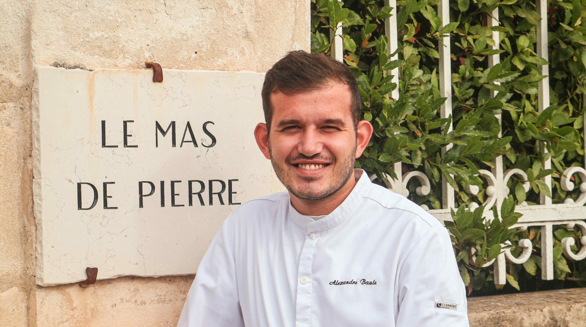 Portrait of Chef Alexandre Baule in front of the sign for the Domaine du Mas de Pierre hotel